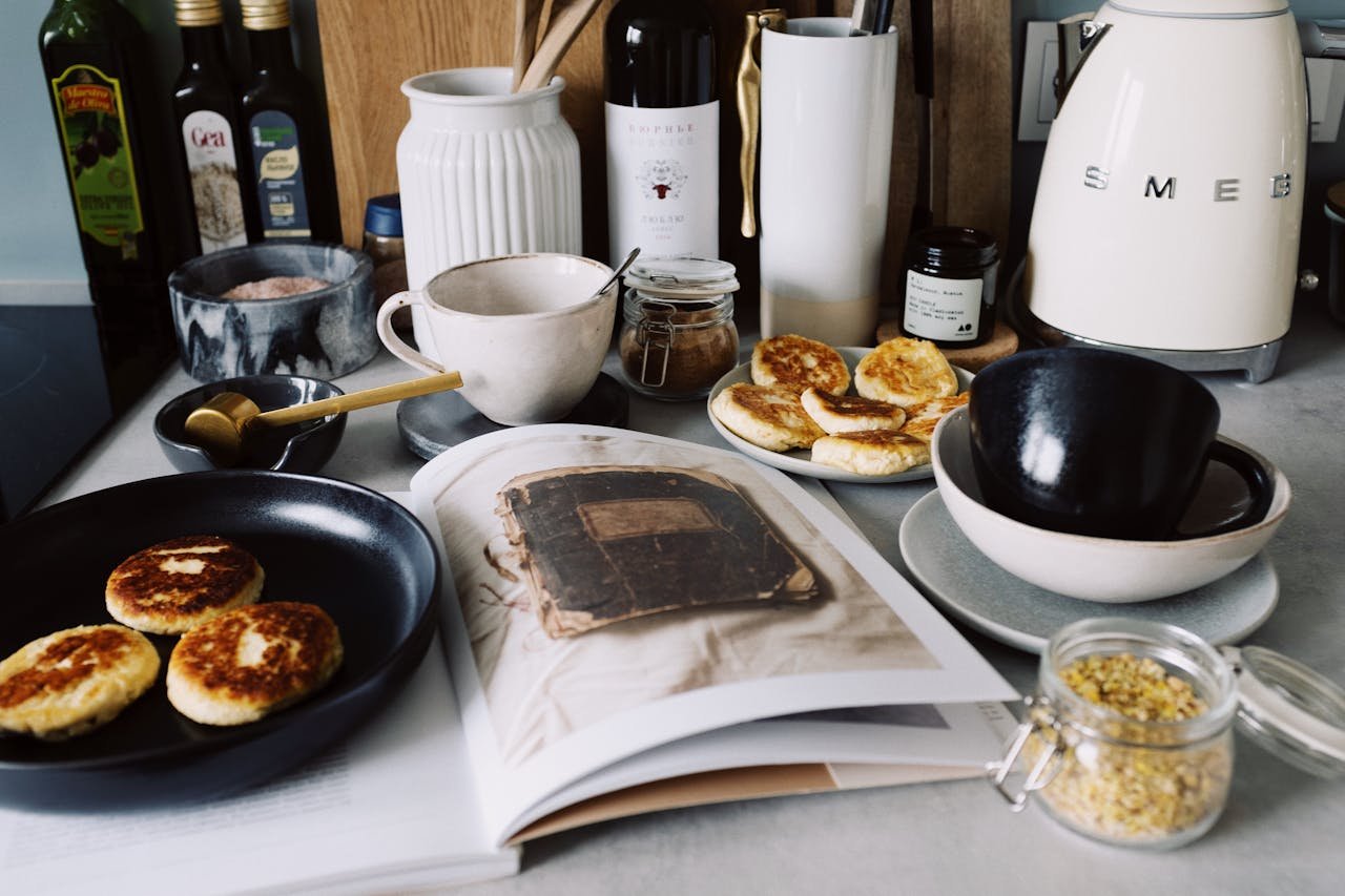 One portion of homemade cheese pancakes in frying pan and another on plate locating near opened stylish magazine among kitchen utensils and appliances in bright kitchen at home